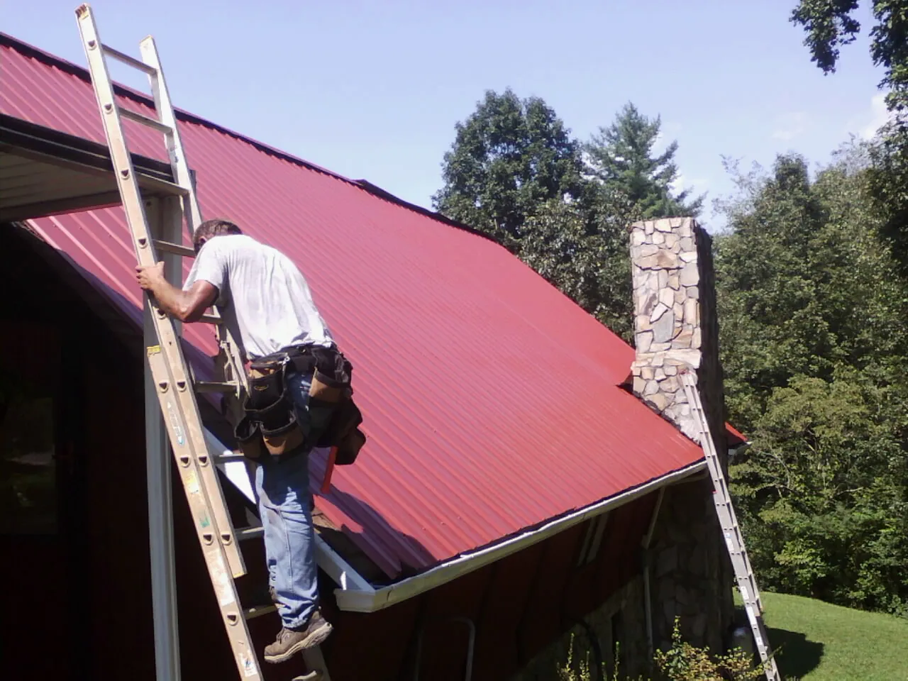 Roofer on a ladder beside a red metal roof and stone chimney — Soffit & Fascia Repair by Roofing Brothers in Newcomerstown